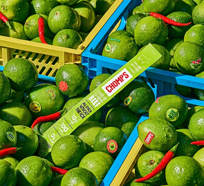 A close-up overhead view of crates filled with bright green limes, with a Chomps Chili Lime Beef stick placed across the crates and a few red chili peppers scattered among the limes.